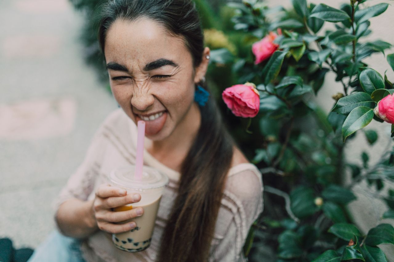 Joyful woman making a funny face while sipping bubble tea beside blooming pink flowers.
